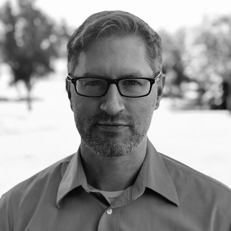 Professional headshot of Patrick Jackson, a man with short light-colored hair and a trimmed beard, wearing glasses and a button-down collared shirt, looking directly at the camera with a calm expression against a softly blurred outdoor bokeh background, in black and white