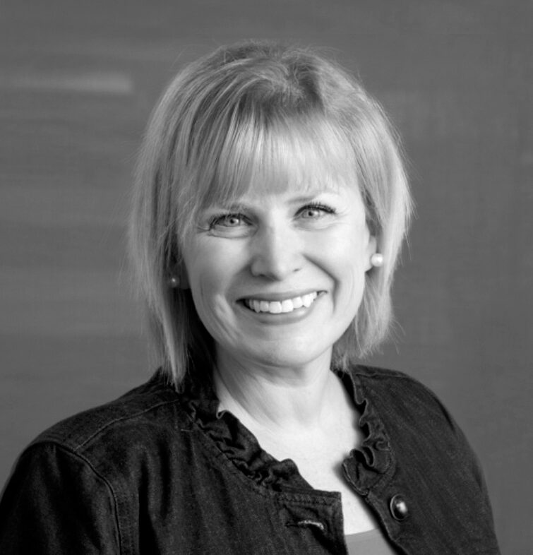 Professional headshot of Kim Gutzwiller, a woman with shoulder-length blonde hair and bangs, smiling warmly at the camera, wearing pearl earrings and a dark ruffled jacket over a lighter top, against a neutral studio background, in black and white