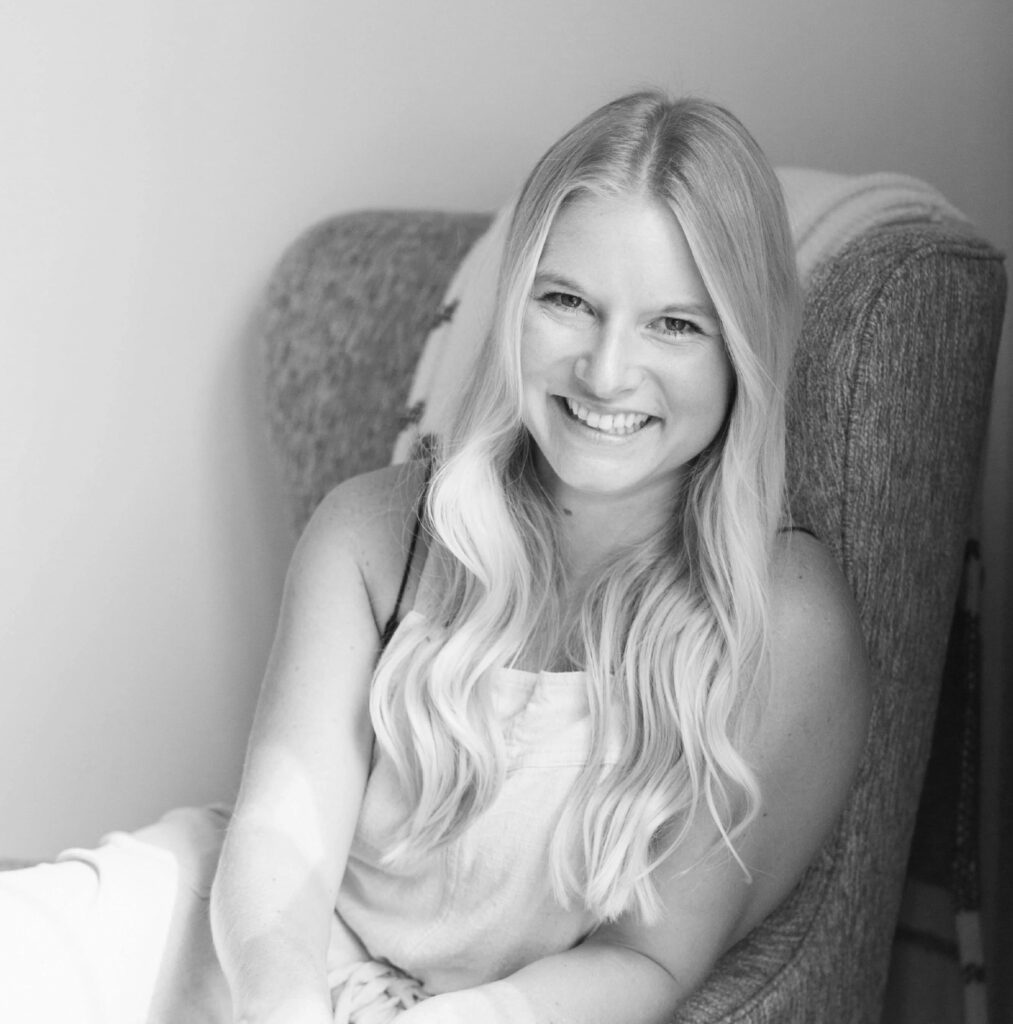 Portrait of Kayla Keith, a young woman with long wavy blonde hair, smiling warmly at the camera while seated and leaning slightly forward on a textured armchair, wearing a sleeveless light-colored top, in black and white