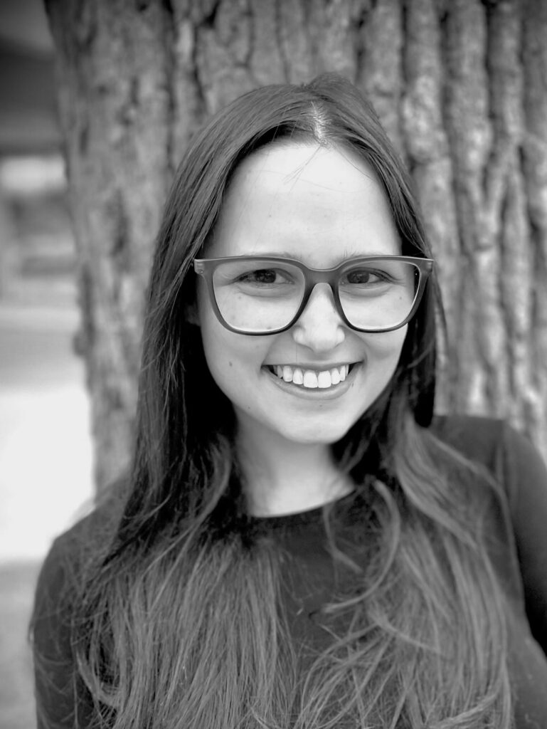 Portrait of Sammi Gassel, a young woman with long straight dark hair, wearing glasses and a dark top, smiling broadly at the camera outdoors with a softly blurred natural background of trees and foliage, in black and white